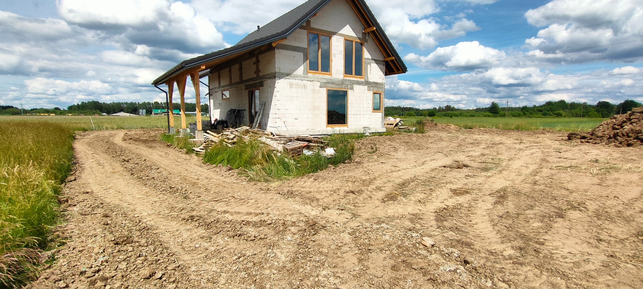 Surowy stan domu z białych bloczków, widoczne drewniane okna i zadaszenie werandy, teren wokół wyrównany, w tle łąka i las.