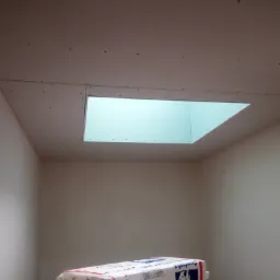 Wnętrze pokoju w trakcie montażu sufitu podwieszanego z płyt kartonowo-gipsowych, widoczne otwór na oświetlenie i paczka materiałów budowlanych na podłodze.