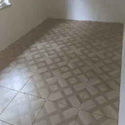 Układanie jasnobrązowej parkietowej mozaiki ceramicznej na podłodze w pustym pokoju, widoczne krzyżyki dystansowe.