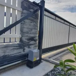 Przesuwna brama wjazdowa z automatyką, widoczny napęd z zębatką, zamontowana na betonowym słupku z elementami dekoracyjnymi.