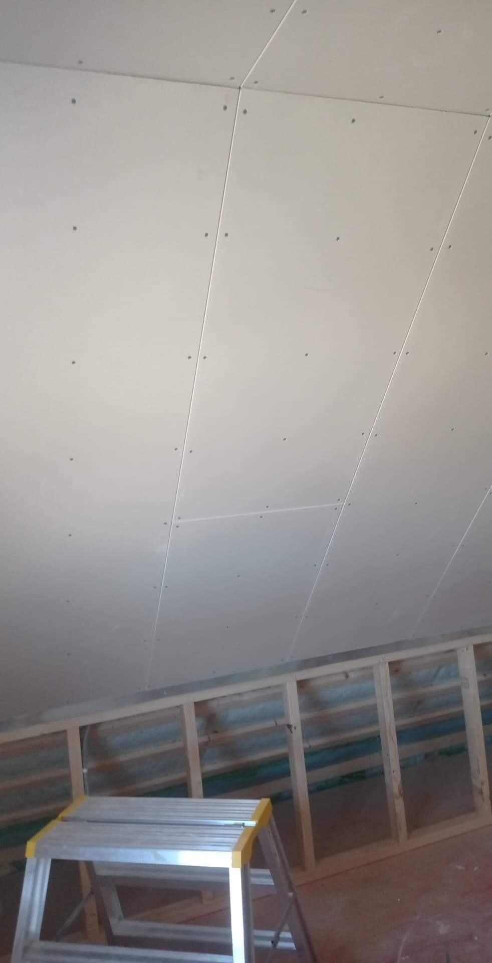 Sufit podwieszany na poddaszu, widoczne łączenia płyt, drewniana konstrukcja nośna i drabina aluminiowa z żółtymi elementami.