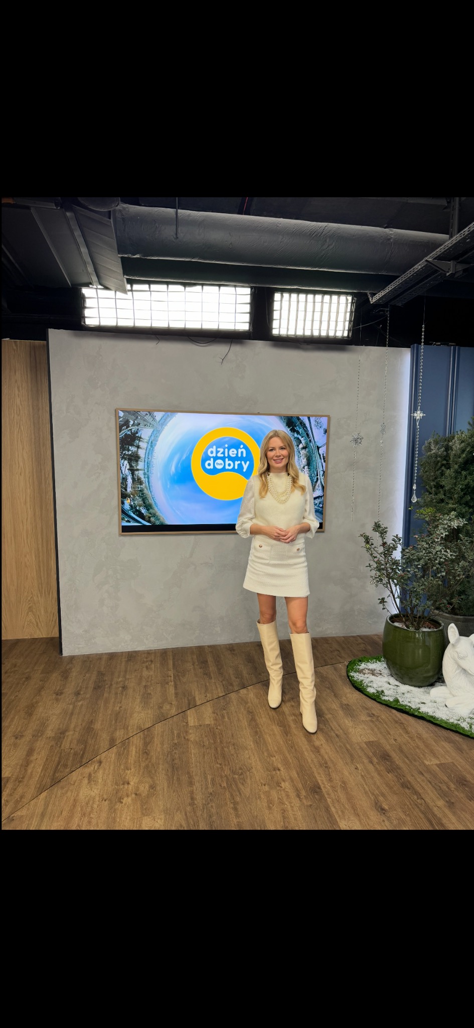 Prezenterka w jasnym stroju na planie studia telewizyjnego z logo 'Dzień Dobry' na ekranie. Oświetlenie studyjne i dekoracje świąteczne w tle.