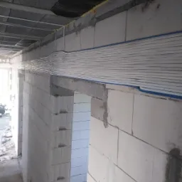 Wiązka przewodów elektrycznych w białej osłonie, poprowadzona wzdłuż ściany z betonu komórkowego w surowym stanie, z widocznymi elementami konstrukcyjnymi stropu i pianką montażową.