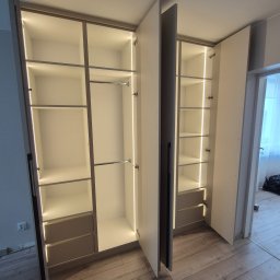 SOCZI MEBLE - Biała szafa wnękowa z oświetleniem LED, widoczne półki i drążki na ubrania. Częściowo otwarte drzwi ukazują wnętrze szafy.