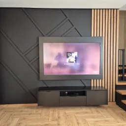 Nowoczesny salon z czarną ścianą akcentową z geometrycznym wzorem i drewnianymi lamelami. Szafka RTV w kolorze grafitowym i drewniana podłoga w jodełkę.
