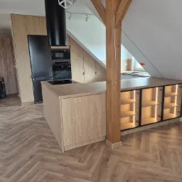 Nowoczesna kuchnia na poddaszu z drewnianymi szafkami, czarnym sprzętem AGD i podświetlaną wyspą. Uwagę zwraca drewniany słup konstrukcyjny.