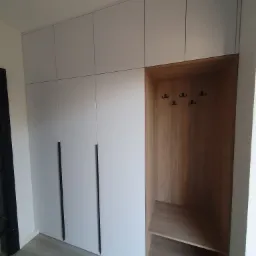 Biała szafa w zabudowie z czarnymi, pionowymi uchwytami, po prawej otwarta wnęka z drewnianym wykończeniem i wieszakami na ubrania.