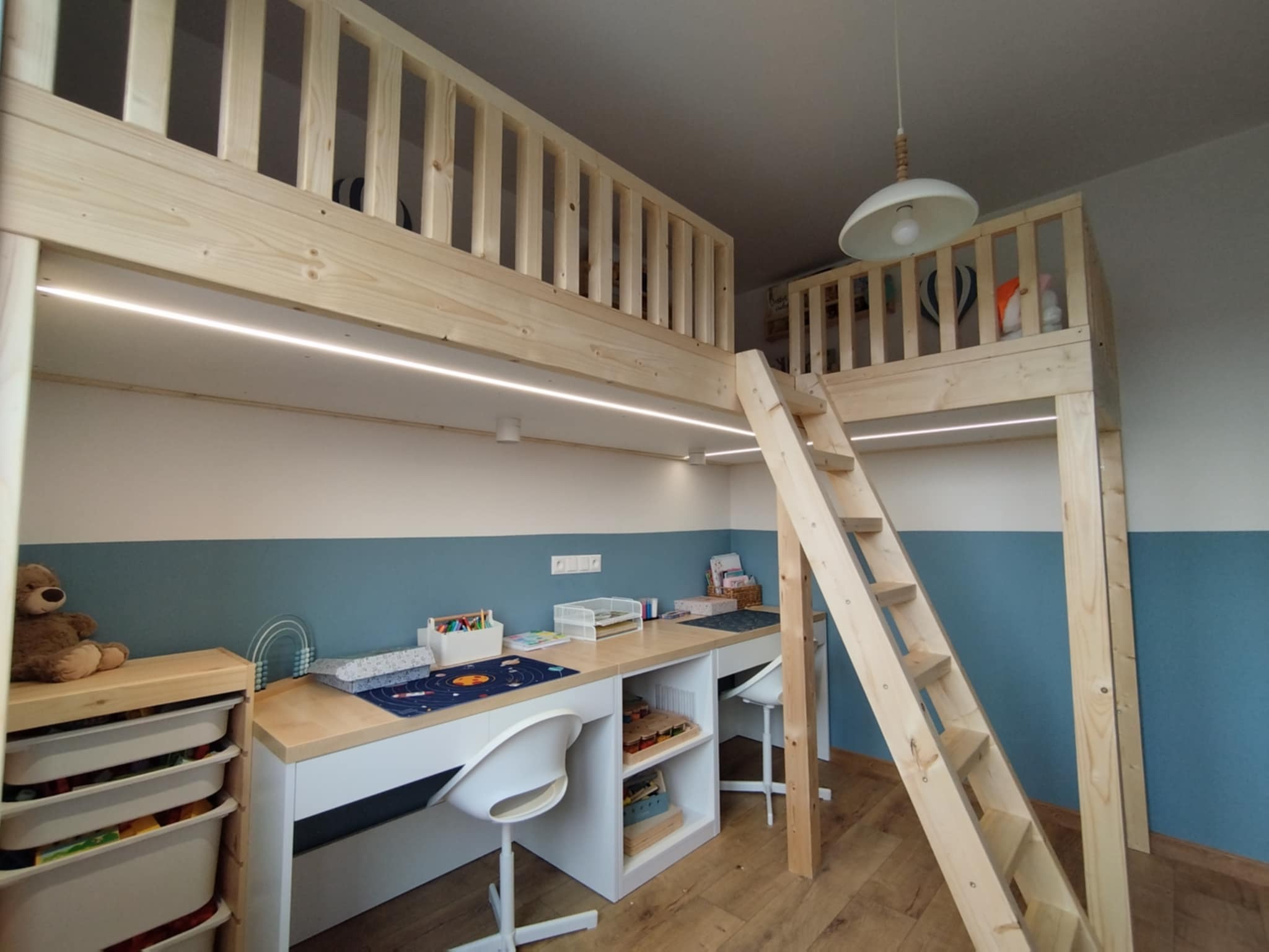 Pokój dziecięcy z drewnianym łóżkiem na antresoli i biurkiem z białymi szafkami. Na biurku akcesoria szkolne, zabawki i lampka. Schody prowadzą na górę.