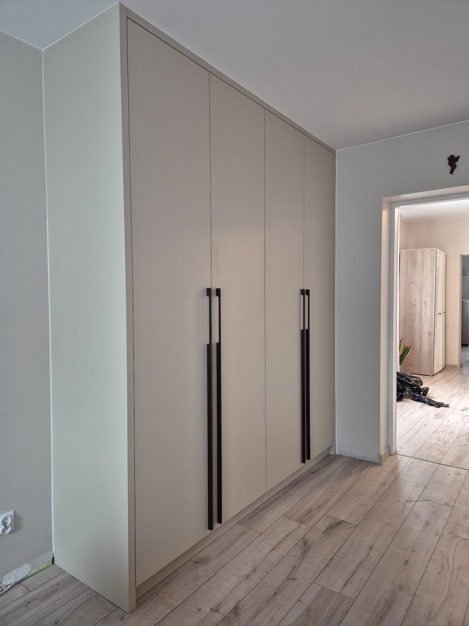 Jasna szafa wnękowa z długimi, czarnymi uchwytami, na tle parkietu i otwartych drzwi do innego pomieszczenia. Minimalistyczny design.