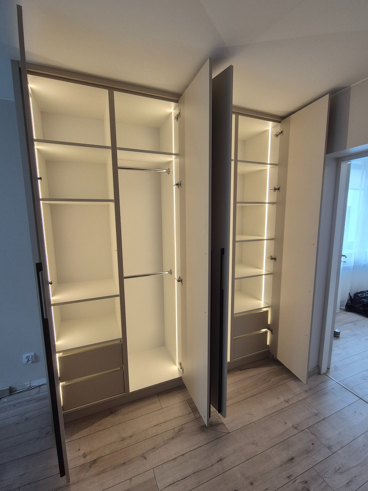 Biała szafa wnękowa z oświetleniem LED, widoczne półki i drążki na ubrania. Częściowo otwarte drzwi ukazują wnętrze szafy.