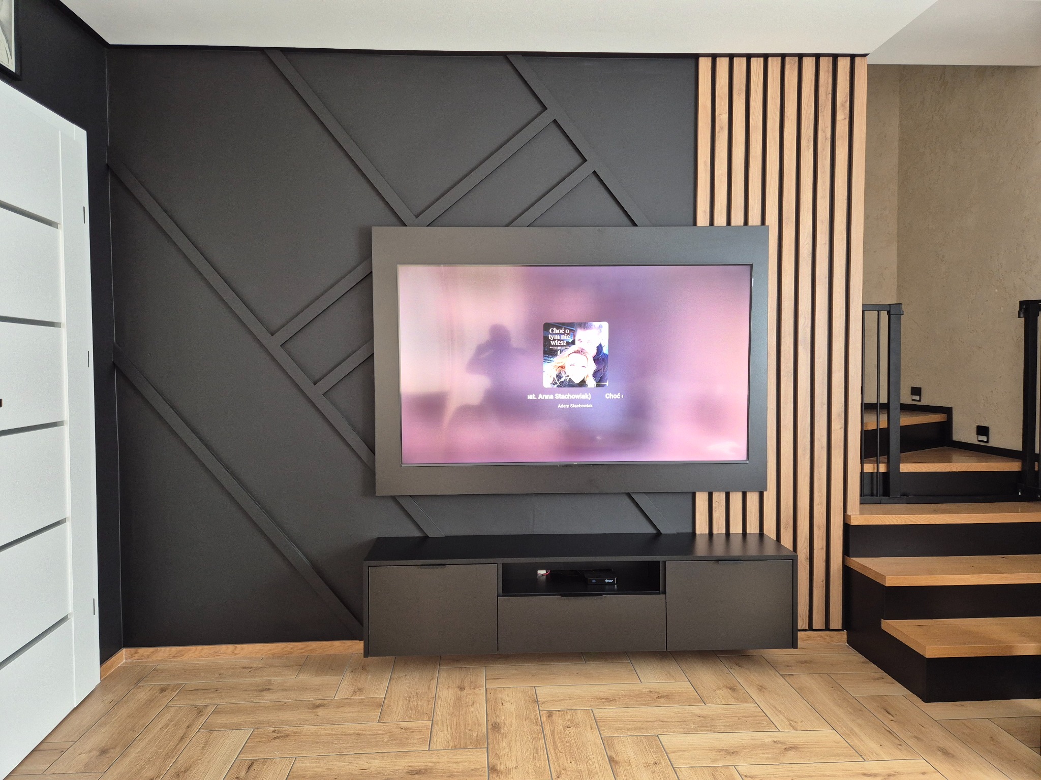 Nowoczesny salon z czarną ścianą akcentową z geometrycznym wzorem i drewnianymi lamelami. Szafka RTV w kolorze grafitowym i drewniana podłoga w jodełkę.