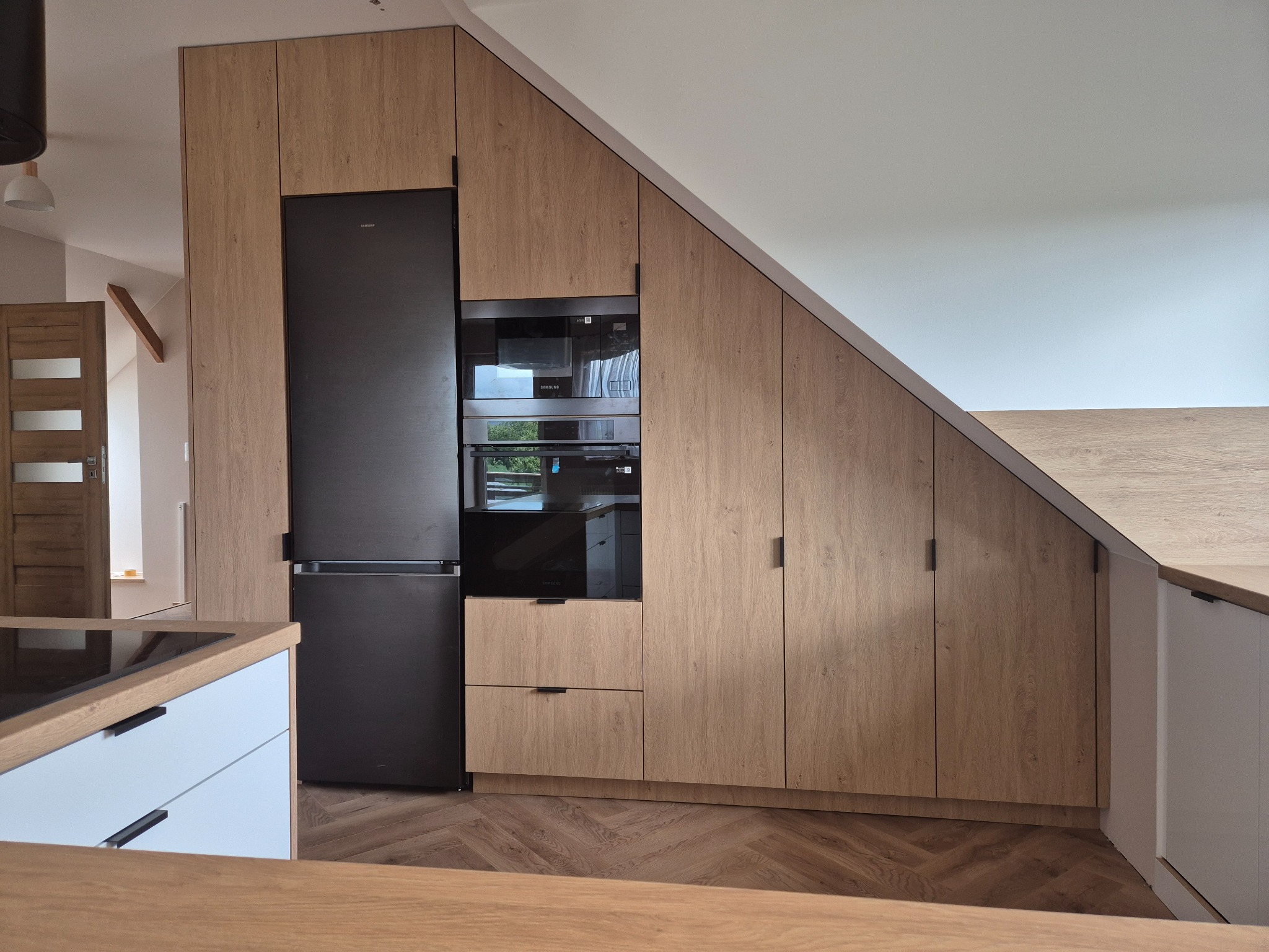 Kuchnia na poddaszu: zabudowa z drewnopodobnymi frontami, czarny sprzęt AGD (lodówka, piekarnik, mikrofalówka). Parkiet w jodełkę na podłodze.