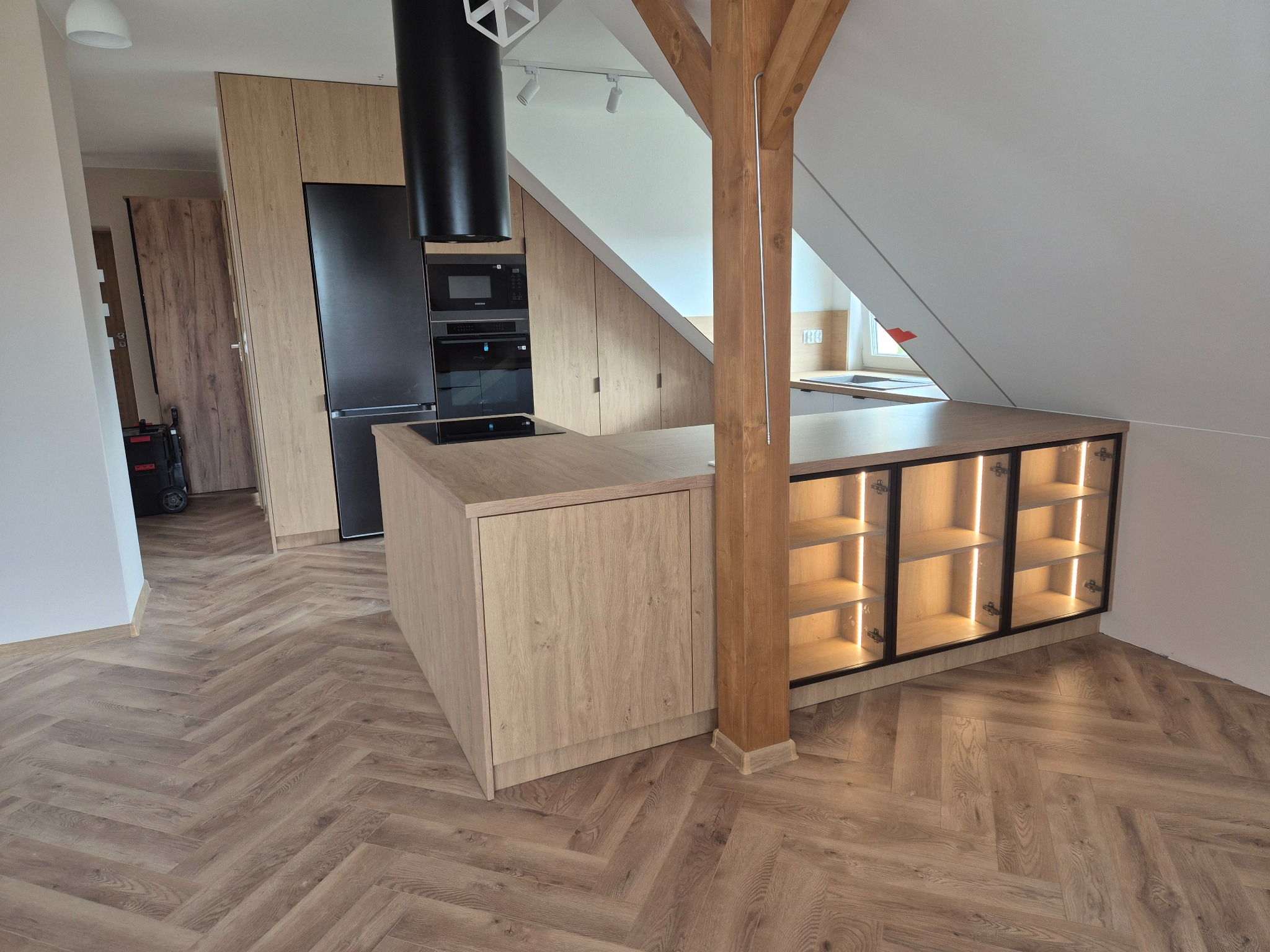 Nowoczesna kuchnia na poddaszu z drewnianymi szafkami, czarnym sprzętem AGD i podświetlaną wyspą. Uwagę zwraca drewniany słup konstrukcyjny.