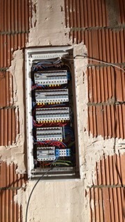 Wnęka w surowej ścianie z czerwonej cegły, w której zamontowana jest biała skrzynka rozdzielcza z zabezpieczeniami i przewodami.