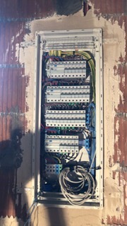 Rozdzielnica elektryczna w trakcie montażu, widoczne przewody i zabezpieczenia, ściana w stanie surowym z widoczną czerwoną cegłą, brakująca obudowa.