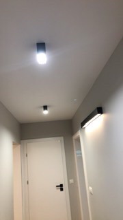 Korytarz z zamontowanym oświetleniem: dwa reflektory sufitowe w czarnej obudowie i podłużna lampa ścienna z białym światłem.