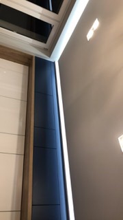 Oświetlenie LED zamontowane wzdłuż ściany i szafki w nowoczesnym wnętrzu, widoczne z perspektywy dolnej, z elementami sufitu i białych drzwi w tle.