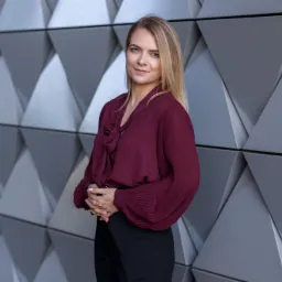 Profesjonalny portret kobiety w bordowej bluzce z kokardą, stojącej na tle nowoczesnej, geometrycznej ściany.