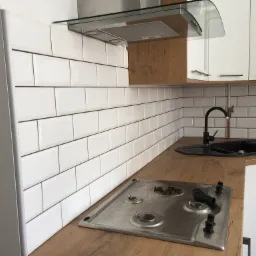 Nowoczesna kuchnia z białymi, prostokątnymi płytkami na ścianie, drewnianym blatem, czarnym zlewem i czarną baterią, oraz stalową płytą gazową z palnikami. W tle białe szafki z przeszklonymi frontami.