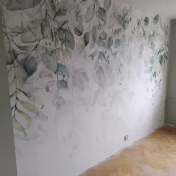 Ściana w pokoju po odświeżeniu z dekoracyjnym motywem akwarelowych liści eukaliptusa, widoczna część drewnianej podłogi w trakcie prac wykończeniowych.
