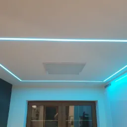 Nowoczesne oświetlenie sufitowe z niebieskimi liniami LED tworzącymi geometryczny wzór, centralny panel oświetleniowy i ciemna zabudowa meblowa w tle.