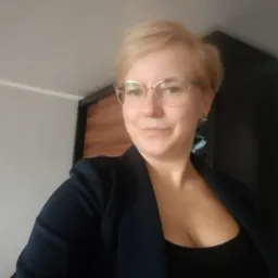 Portret kobiety w okularach, z krótkimi blond włosami, ubranej w czarną bluzkę i granatową marynarkę, uśmiechającej się do kamery na tle ściany i ciemnych drzwi.