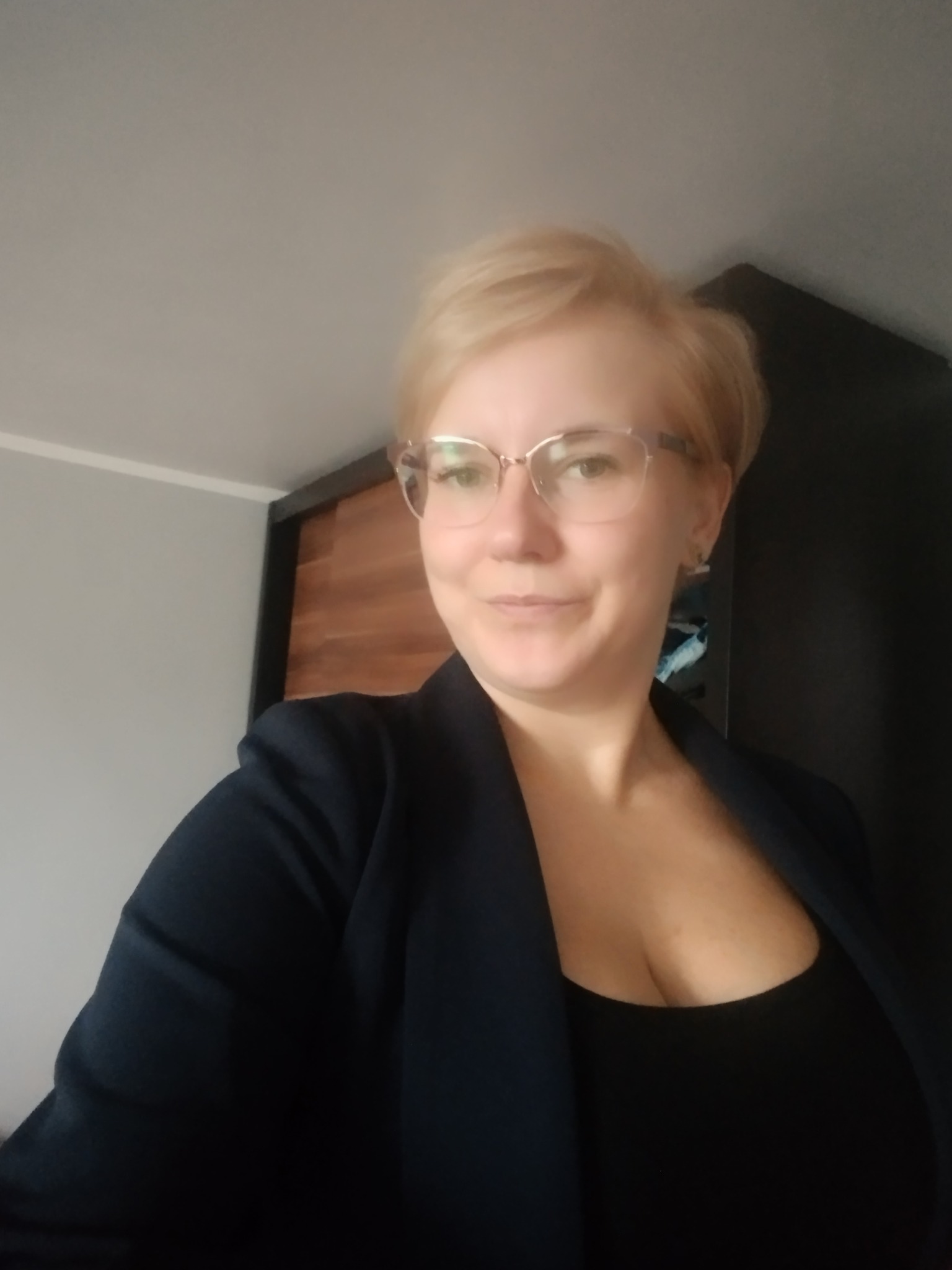 Portret kobiety w okularach, z krótkimi blond włosami, ubranej w czarną bluzkę i granatową marynarkę, uśmiechającej się do kamery na tle ściany i ciemnych drzwi.