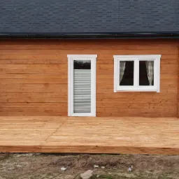 Dom z drewnianą elewacją i nowym tarasem, widoczne elementy konstrukcyjne tarasu w trawie, okna z białymi ramami i grafitowy dach.