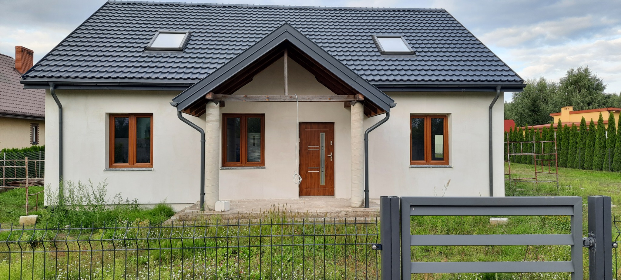 Ocieplony dom jednorodzinny z ciemną dachówką i drewnianymi oknami, widoczny zza metalowego ogrodzenia, z fragmentem sąsiedniego budynku w tle.