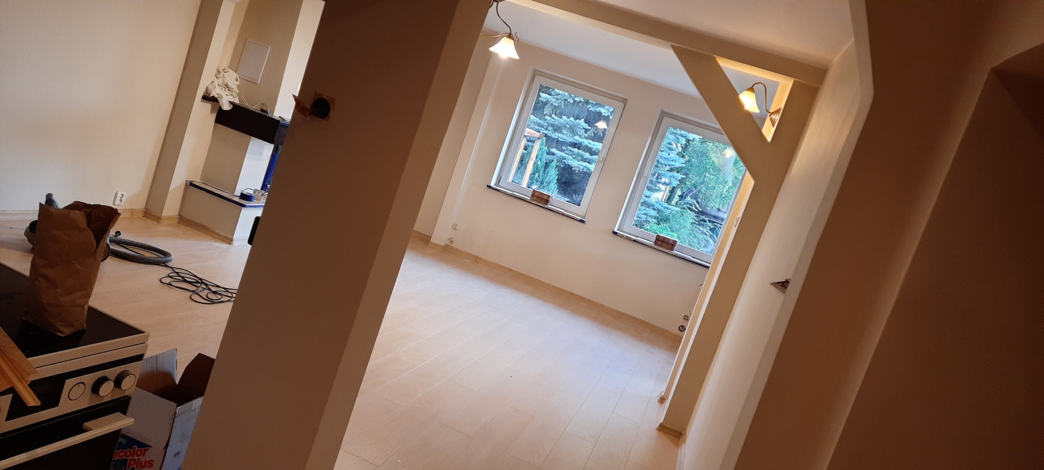 Wnętrze w trakcie wykańczania: świeżo położona podłoga z paneli, pomalowane ściany, widoczne gniazdka elektryczne, przez okna widać drzewa, w tle kominek, narzędzia i materiały budowlane.