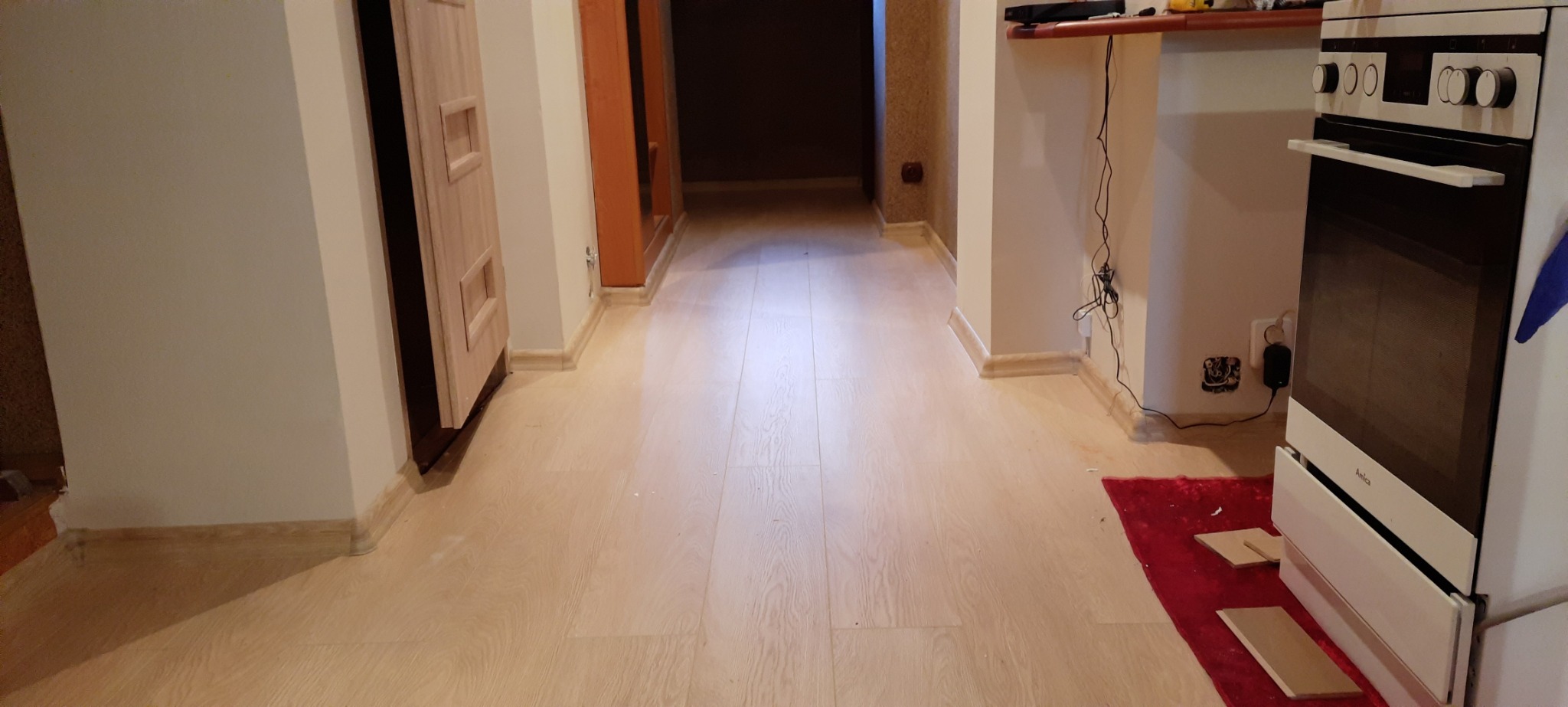 Jasne panele podłogowe w korytarzu, widoczne listwy przypodłogowe, fragment kuchni z kuchenką Amica i czerwonym dywanikiem.