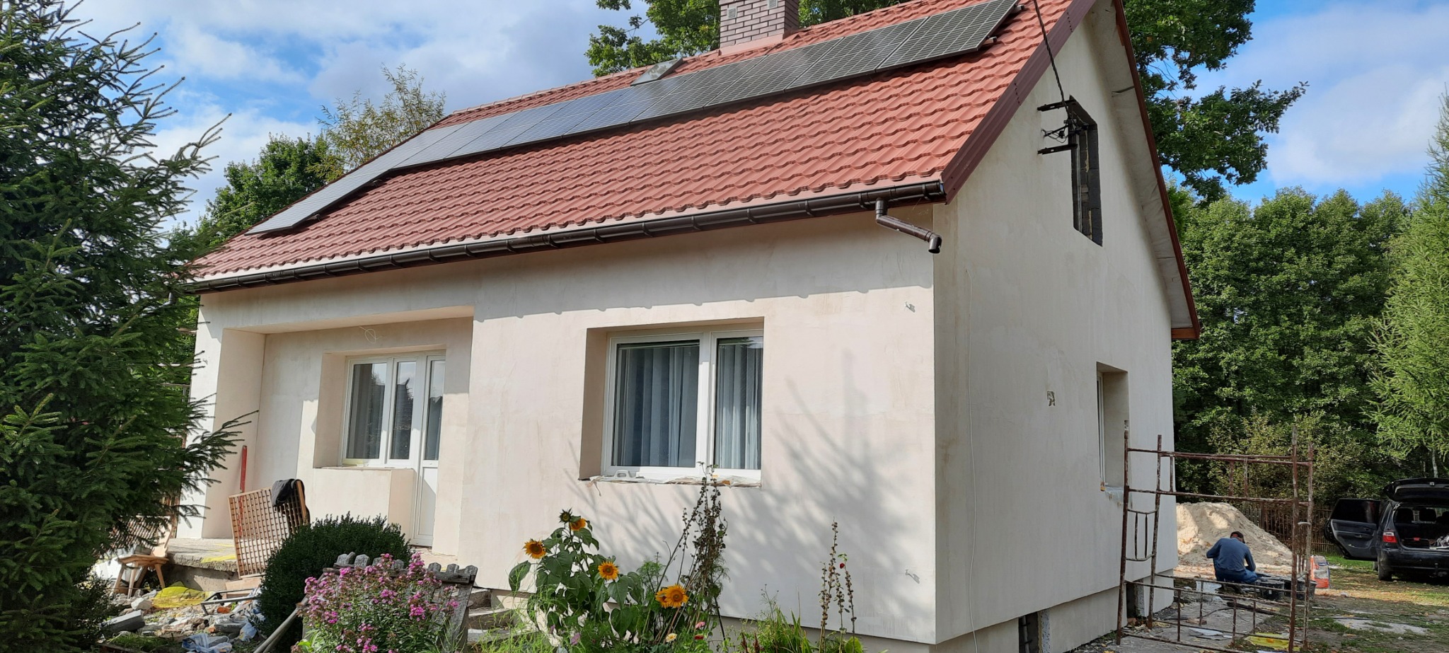 Dom jednorodzinny z nałożoną warstwą szpachli na elewacji, z widocznymi panelami słonecznymi na dachu i pracownikiem przygotowującym narzędzia na rusztowaniu obok samochodu z otwartym bagażnikiem.