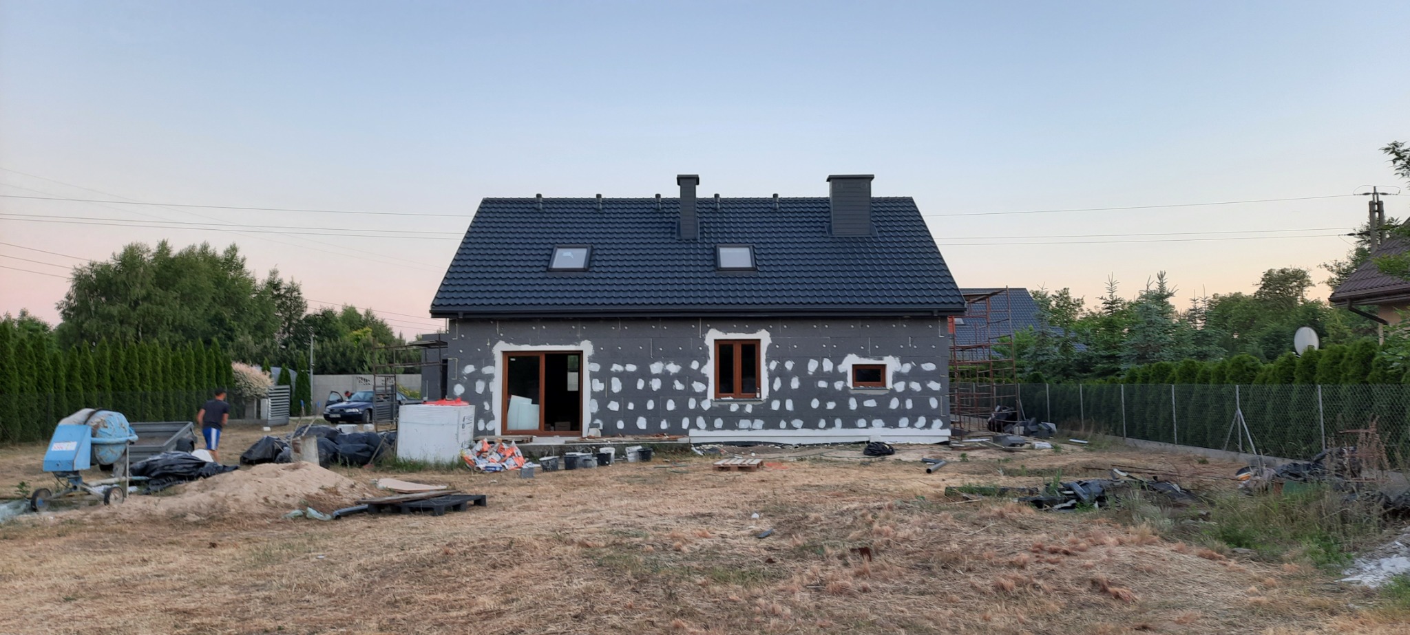 Dom jednorodzinny w trakcie ocieplania elewacji, widoczne szare płyty izolacyjne z punktowo nałożoną zaprawą, brązowe ramy okienne, ciemnoszara dachówka, w tle betoniarka i osoba.