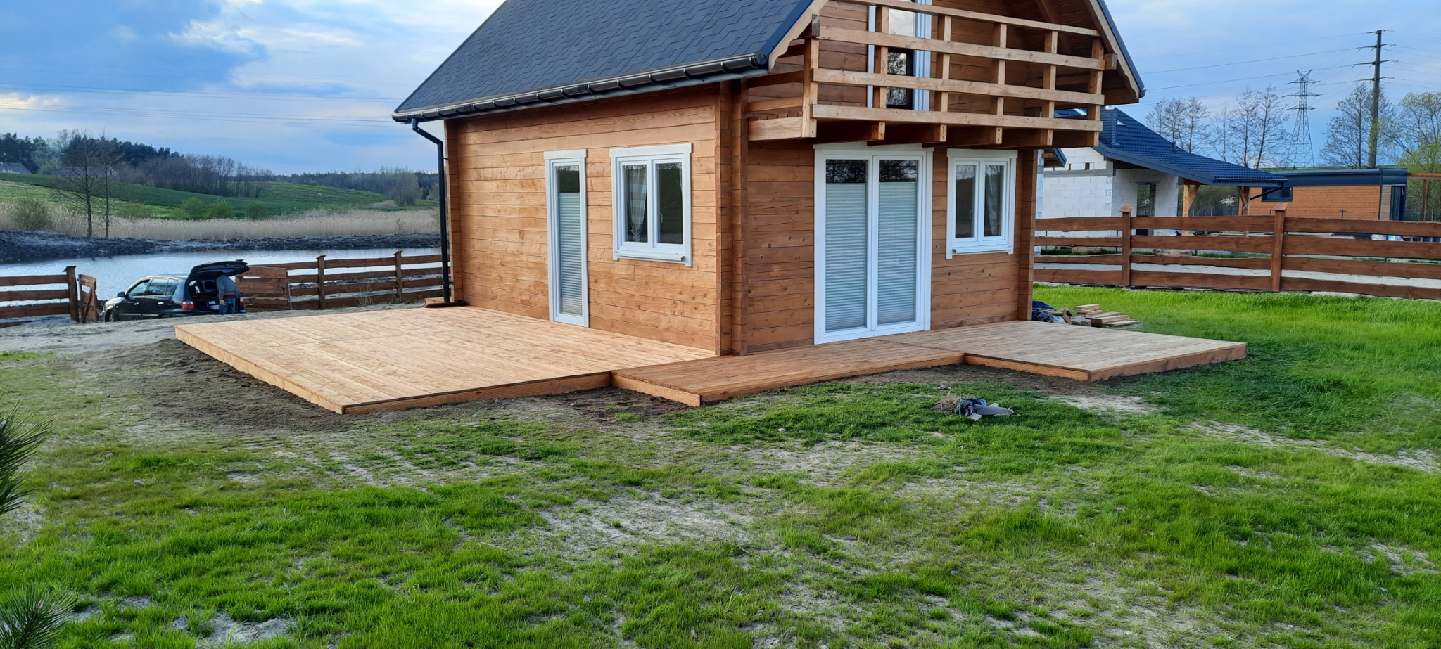 Drewniany domek letniskowy z nowo wybudowanym tarasem, widok na jezioro, w tle samochód z otwartym bagażnikiem.