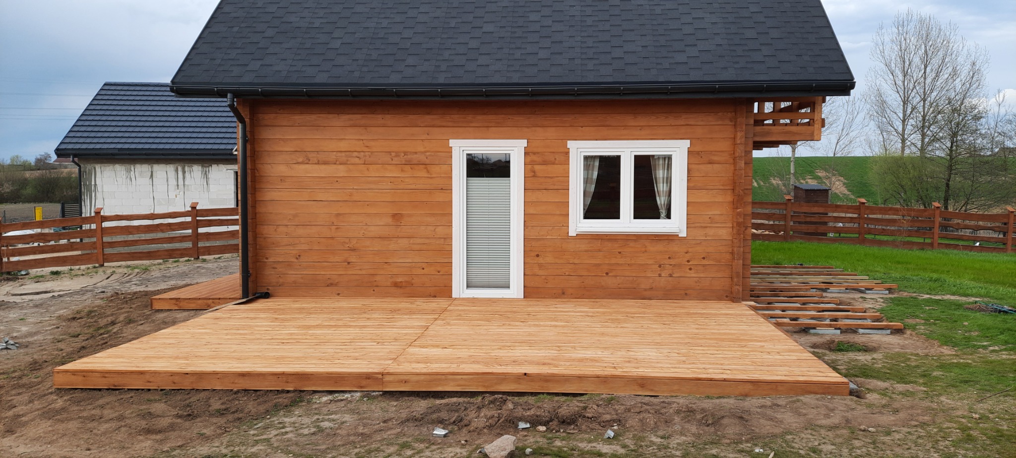 Dom z drewnianą elewacją i nowym tarasem, widoczne elementy konstrukcyjne tarasu w trawie, okna z białymi ramami i grafitowy dach.