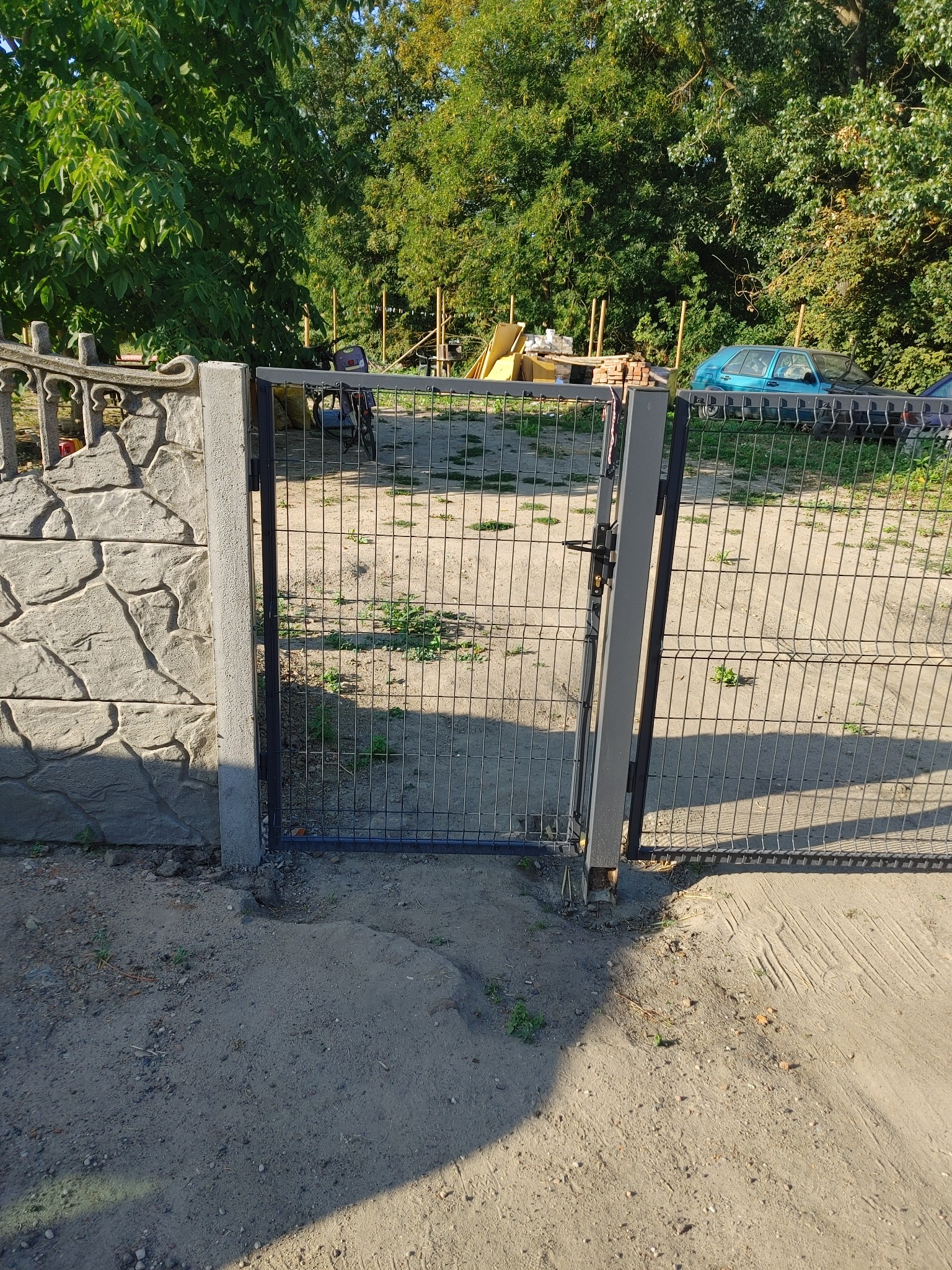 Otwarta metalowa furtka z zamkiem, osadzona między betonowym słupkiem z imitacją kamienia a metalowym słupkiem, prowadząca na teren z piaskiem, trawą, rowerem, stertą desek i zaparkowanym...