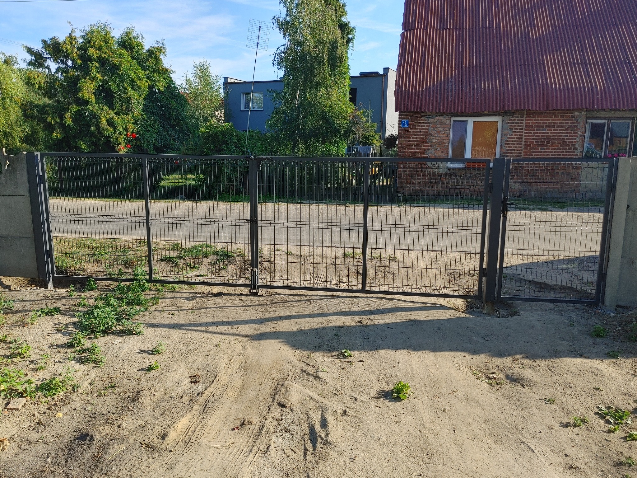 Nowoczesna, metalowa brama wjazdowa z siatki, z widocznymi śladami opon na piaszczystym podłożu, prowadząca do posesji z domem o ceglanej elewacji i czerwonym dachem.
