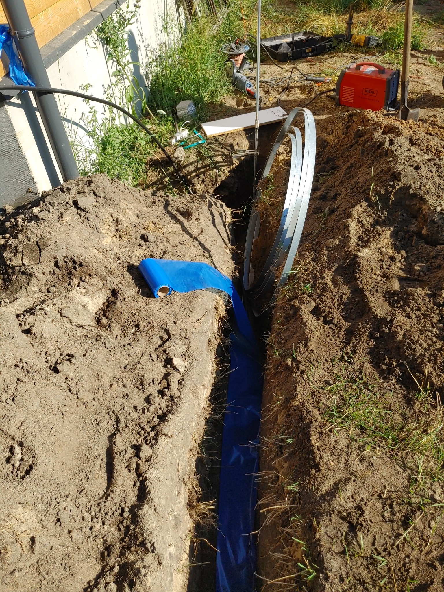 Wykop w ziemi wyłożony niebieską folią, obok leżą metalowe rurki, w tle widoczny agregat spawalniczy i narzędzia.