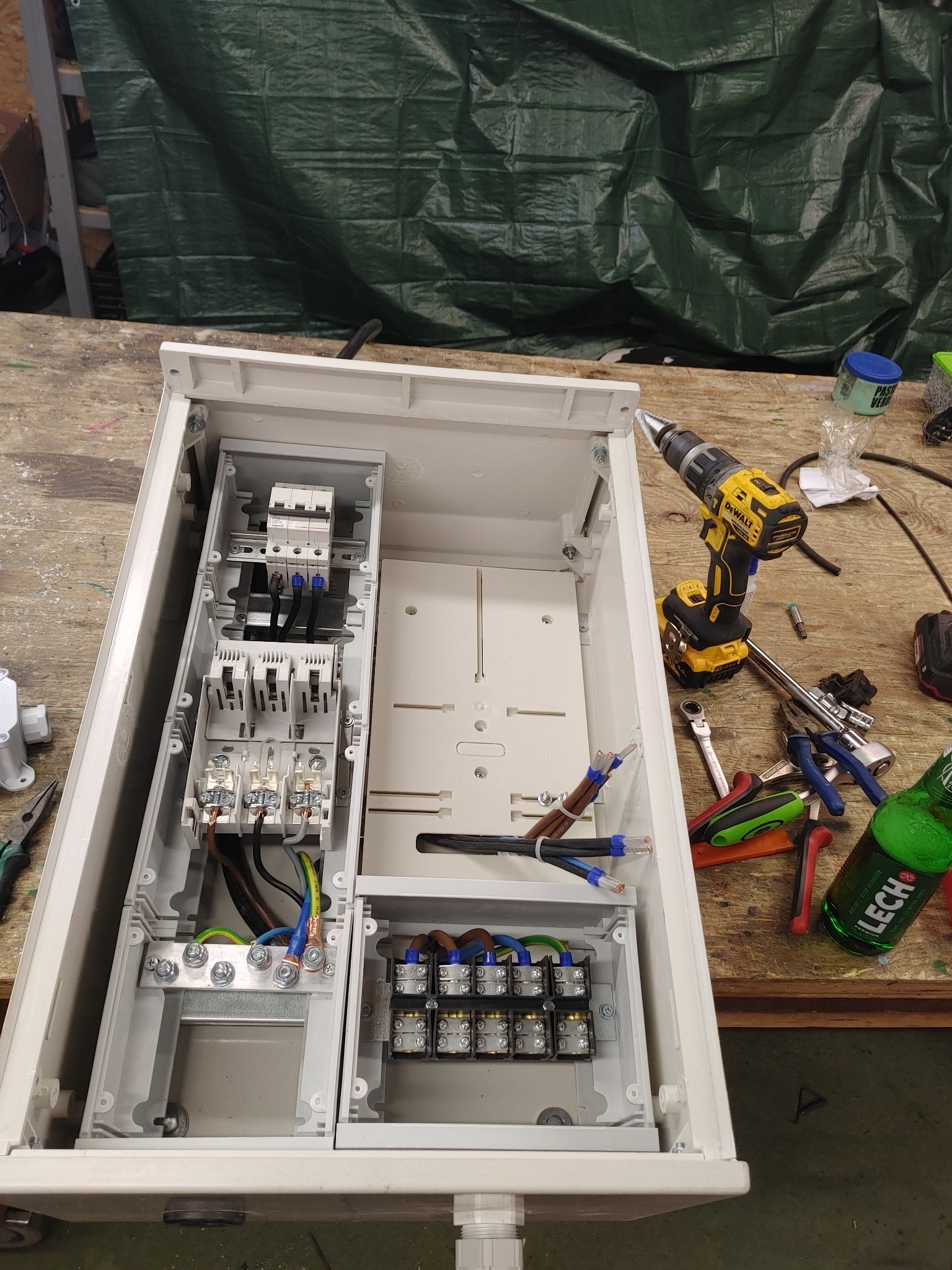 Otwarta biała skrzynka elektryczna w trakcie montażu z widocznymi przewodami, bezpiecznikami i listwami zaciskowymi, obok wkrętarka DeWalt, narzędzia i butelka piwa Lech.