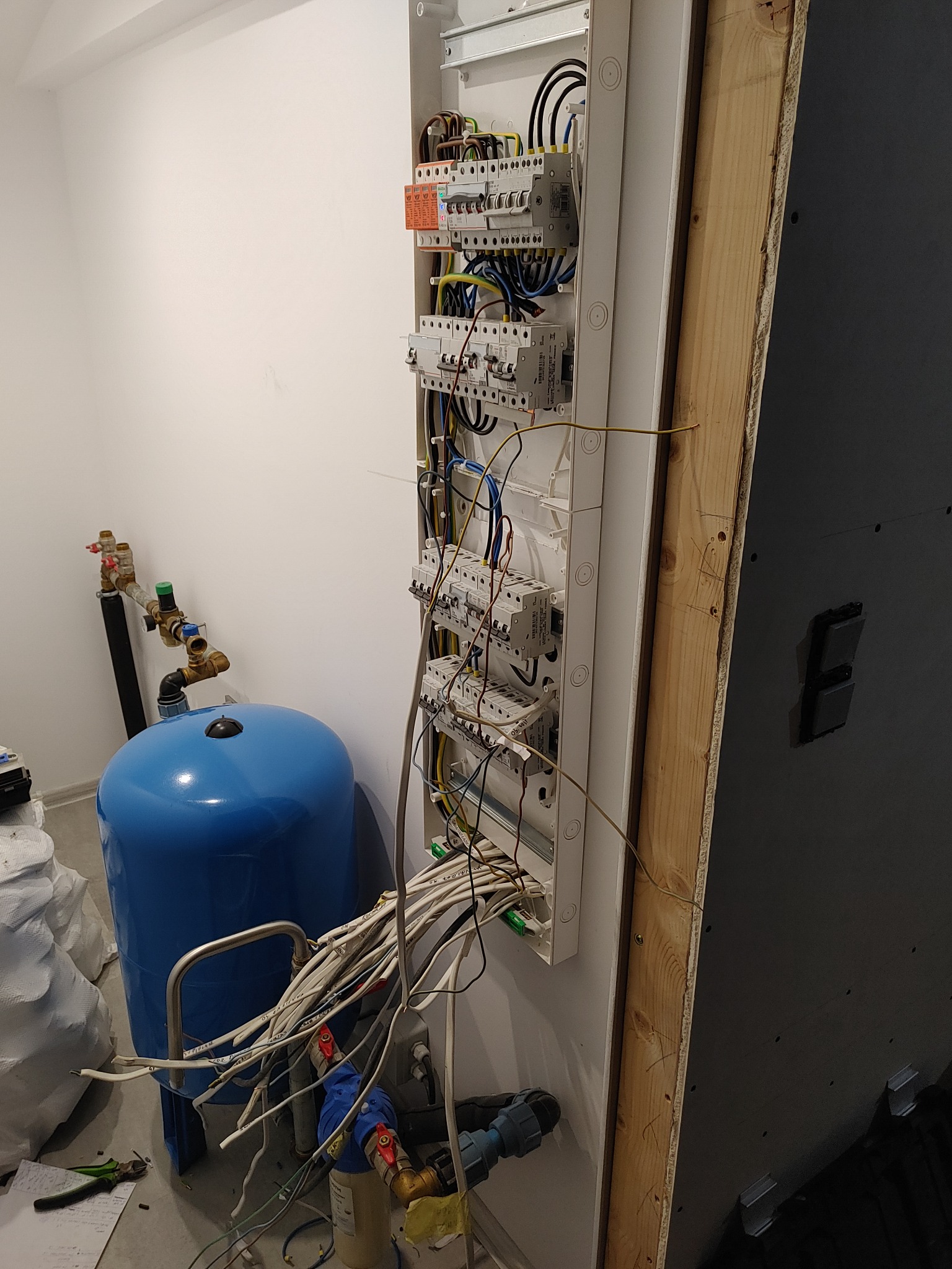 Rozdzielnica elektryczna z widocznymi przewodami, zamontowana na ścianie w pomieszczeniu technicznym, obok niebieski zbiornik hydroforowy i fragment ściany z płyt OSB.