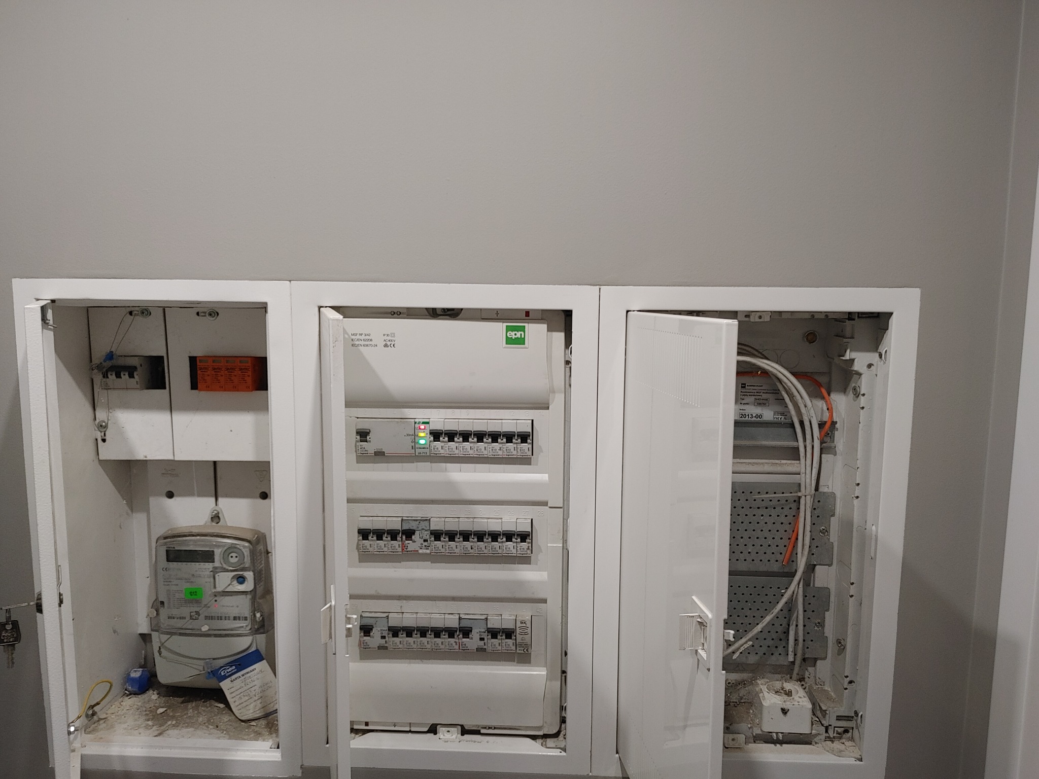 Otwarta szafka elektryczna z licznikiem energii po lewej, rozdzielnicą elektryczną w środku i przestrzenią na przewody po prawej stronie, widoczne elementy instalacji elektrycznej.