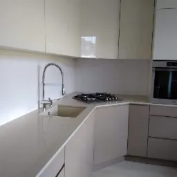 Jasna, minimalistyczna kuchnia na wymiar z beżowymi frontami szafek, chromowaną baterią z wyciąganą wylewką i płytą gazową na blacie.