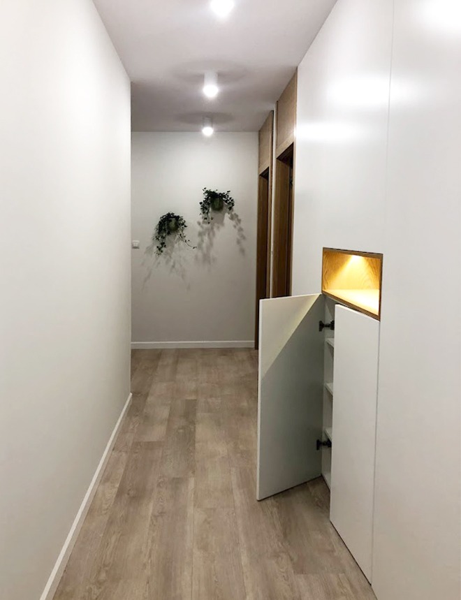Wnętrze korytarza z białą zabudową meblową z oświetloną wnęką, otwartymi drzwiczkami szafki i drewnianą podłogą, dwie wiszące rośliny na ścianie w tle.