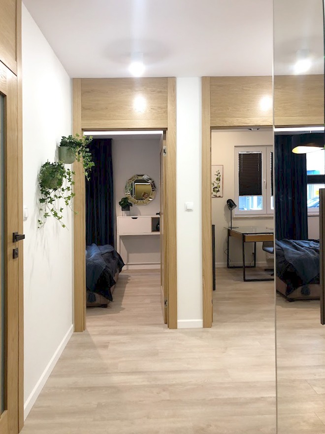 Korytarz z jasną podłogą i dwiema parami drzwi z drewnianymi ościeżnicami, widok na pokoje z granatowymi zasłonami, lustro na ścianie odbija widok pokoju