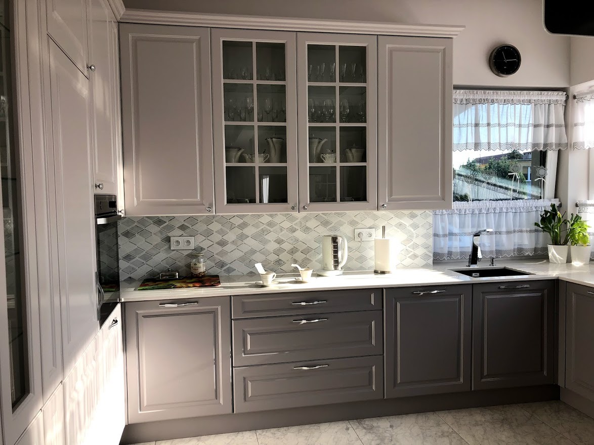 Jasna kuchnia w stylu klasycznym z szarymi frontami, szklanymi witrynami górnych szafek, mozaiką na ścianie nad blatem i widokiem z okna na zieleń.
