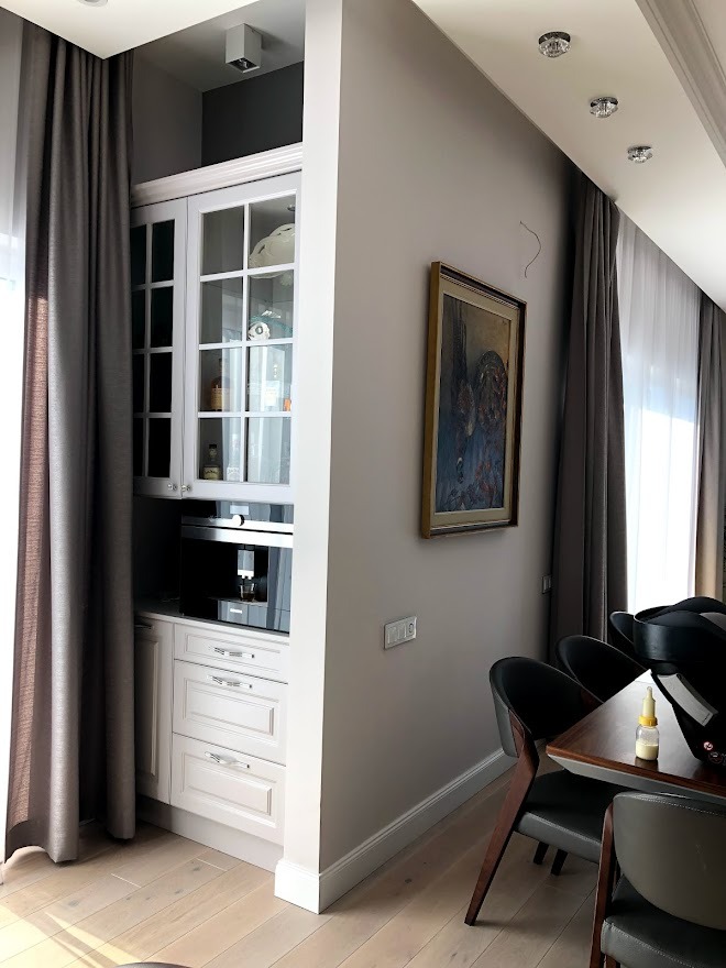 Biała zabudowa kuchenna z przeszkloną szafką i wbudowanym ekspresem do kawy, uzupełniona wiszącym obrazem w klasycznej ramie w nowoczesnym wnętrzu z drewnianą podłogą i szarymi zasłonami.