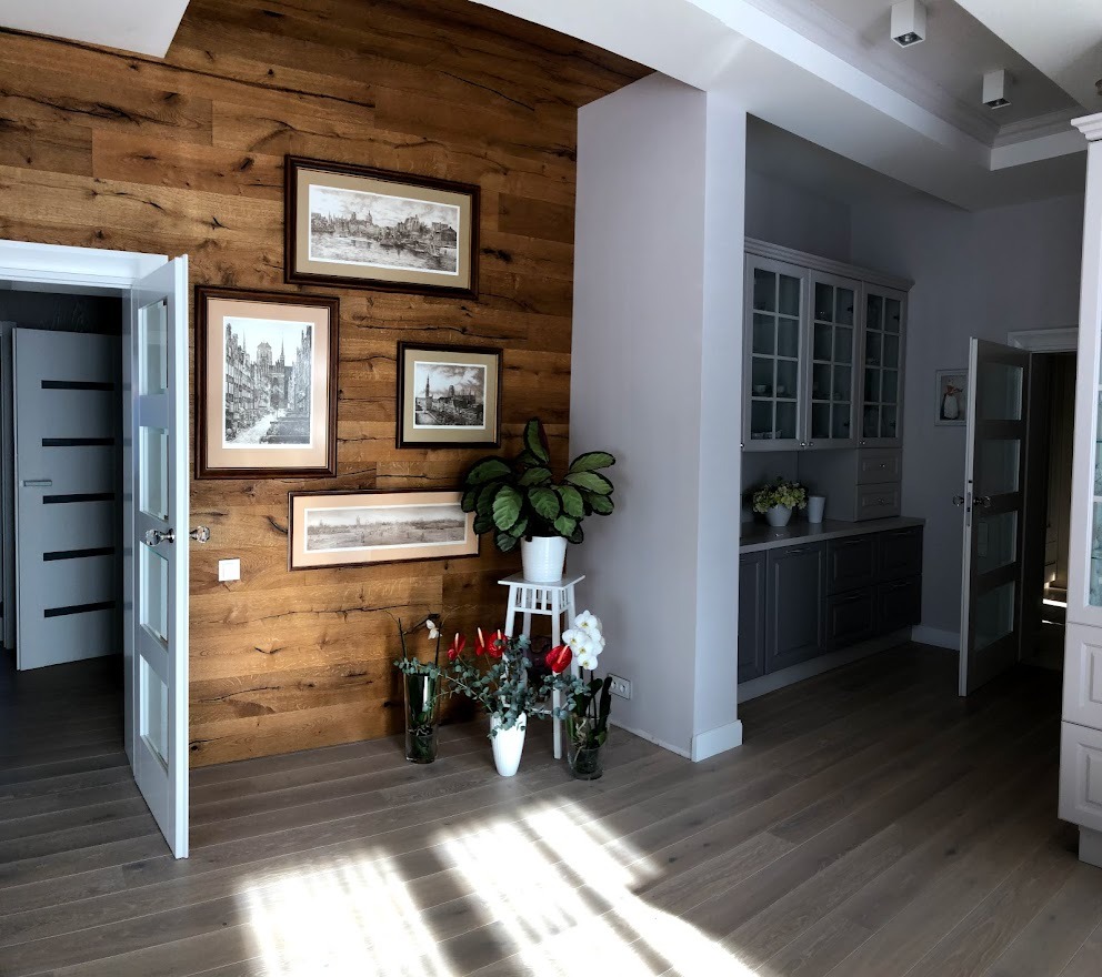 Salon z drewnianą ścianą, dekoracjami w postaci obrazów i roślin, oraz widokiem na kuchnię z szarymi szafkami i przeszklonymi witrynami.