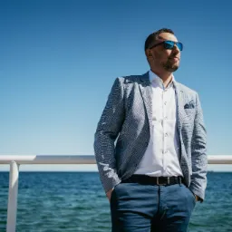 Mężczyzna w marynarce w kratę i okularach przeciwsłonecznych, stojący na tle morza i błękitnego nieba, z rękami w kieszeniach spodni.
