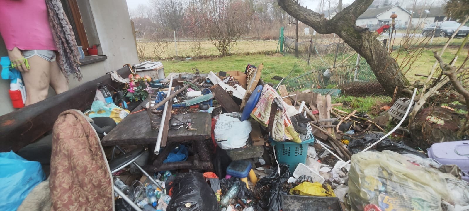 Sterta śmieci i odpadów na zewnątrz budynku, widoczne meble, worki, plastikowe pojemniki i inne przedmioty, sugerujące potrzebę opróżniania mieszkania lub wywozu gruzu.