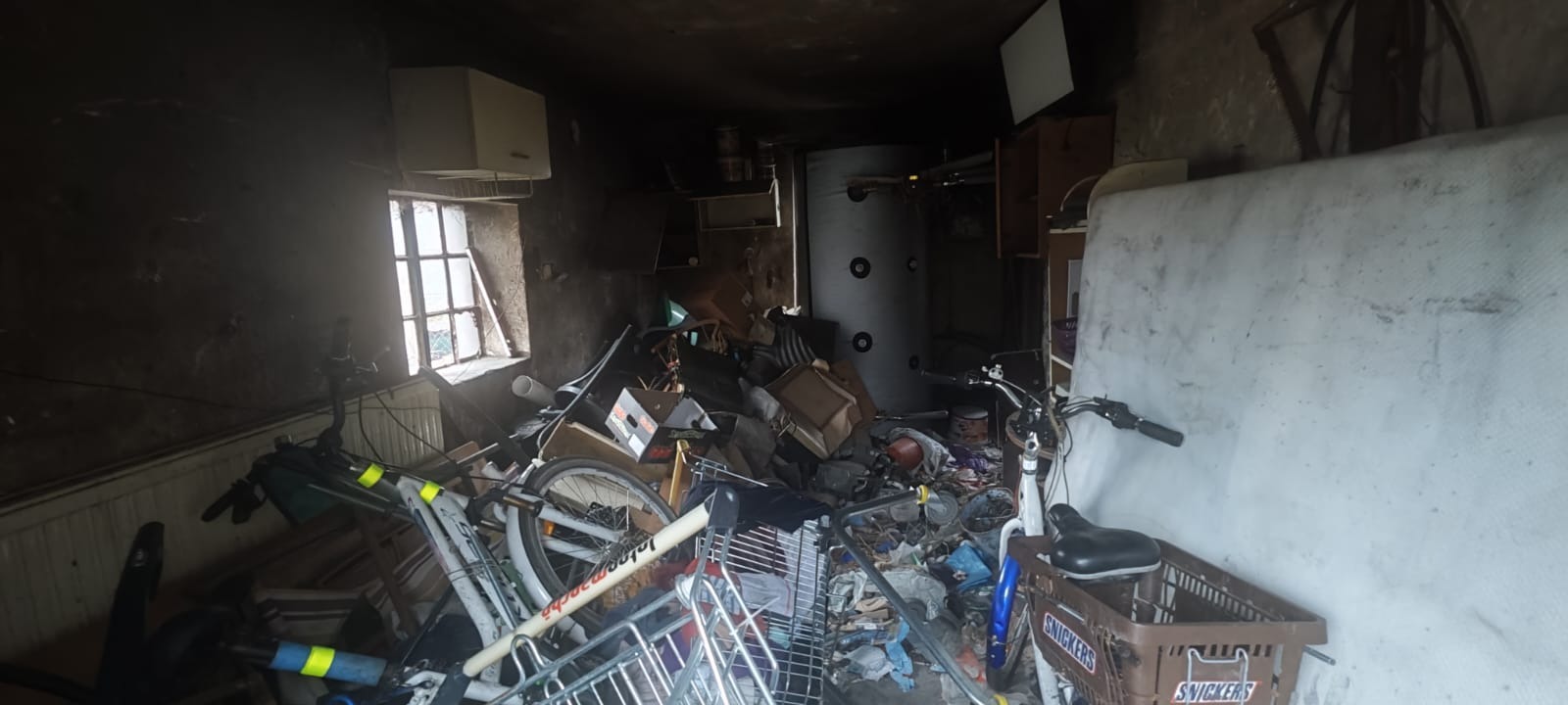 Zagracone pomieszczenie z oknem, wypełnione różnymi przedmiotami, w tym rowerami, kartonami i materacem. Widoczny bojler i kosz z logo Snickers.